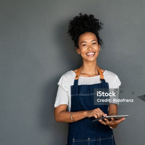 Portrait of beautiful young cafe black waitress using digital tablet on grey background. Happy young black woman in blue jeans apron working on digital tablet while looking at camera. Successful african american cafe owner isolated against gray wall with copy space.